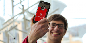 Student indoors at the Engineering Centers Building holding a cell phone with a phone wallet attached. The wallet displays the College of Engineering logo. Camp Randall Stadium is visible through a window in the background.